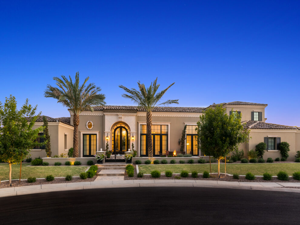 A large, modern luxury home with a landscaped front yard, palm trees, and pathway lighting at dusk.
