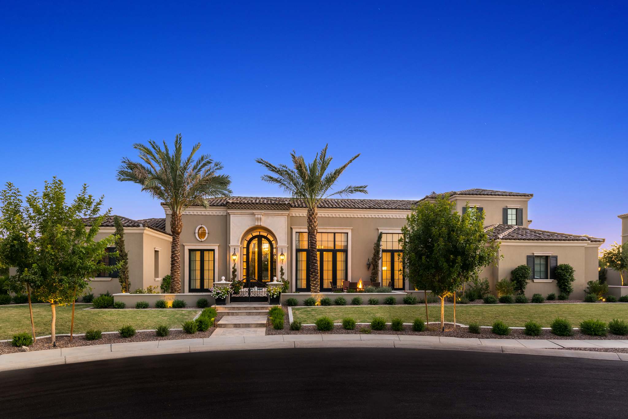 A large, modern luxury home with a landscaped front yard, palm trees, and pathway lighting at dusk.