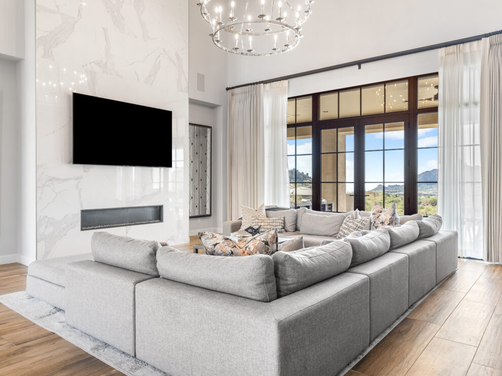 Living Room: A modern living room with a large sectional grey sofa, a flat-screen TV mounted on a marble wall, and a large chandelier hanging from the ceiling. The room features floor-to-ceiling windows with white curtains that offer a view of the outdoors.